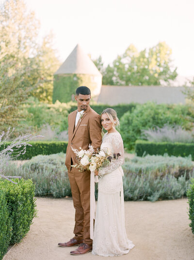 groom in a light brown suit looking back and down at his bride holding a bouquet resting her head behind him on his shoulder