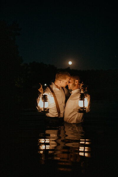 Grooms kissing in the moonlight while holding lanterns and standing in the water