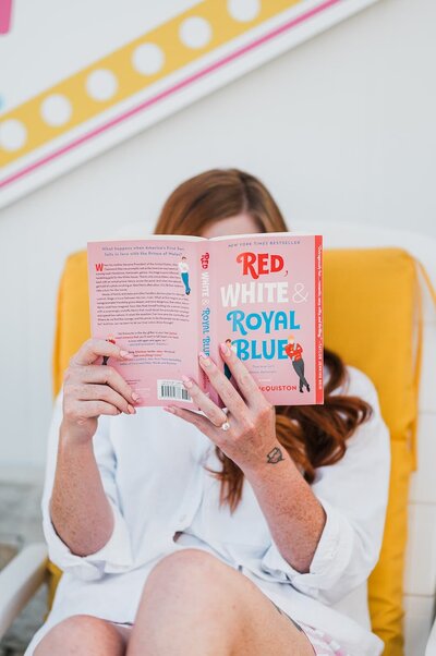 A woman with red hair reads “Red, White & Royal Blue” while lounging on a yellow chair, embodying a playful and relaxed moment.