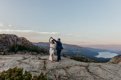 A couple enjoys a romantic Lake Tahoe elopement, sitting on a blanket by the sandy shore, toasting drinks as the sun sets. The woman wears a blue and white striped dress, and the man wears a white shirt. Snow-capped mountains and the lake’s clear waters create a stunning backdrop.