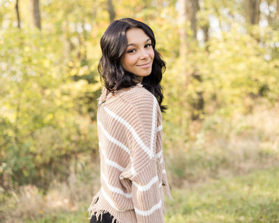 high school female senior, with long black hair, standing in grass with a tree line next to her, standing to the side, looking at the camera smiling softly, wearing black leggings, and light tan and white stripped shirt