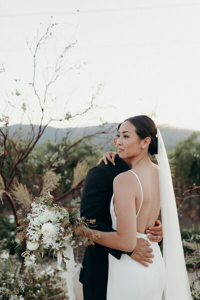 Bride with groom, holding her white bouquet, posing in a California vineyard