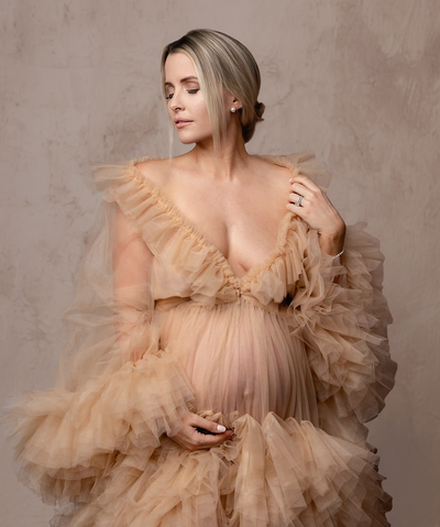 Pregnant mother wearing a sheer champagne colored ruffled dress that shows her baby bump.