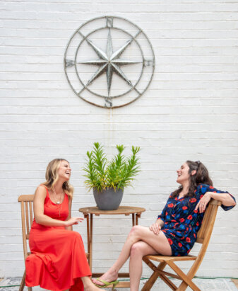 two white women wearing sundresses sit at a table outdoors under a large metal direction compass laughing