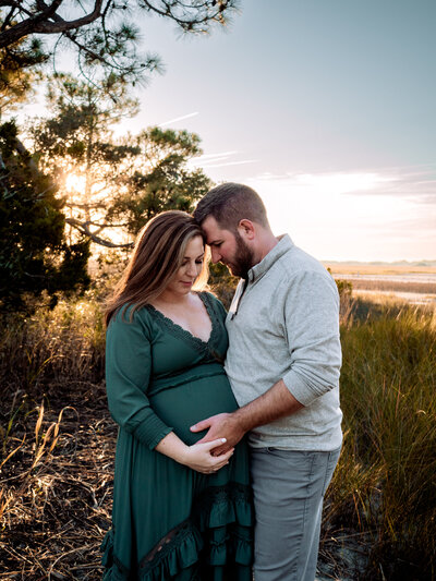 A couple embraces, looking down at the woman's pregnant belly. The husband holds her pregnant belly. They are standing on the marsh with tall grass and trees behind them, and the sun shining through the trees.