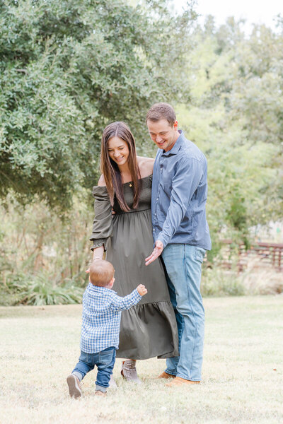 A couple stands on grass in front of a bridge at Katherine Fleischer Park. Their baby boy runs toward them, The parents are smiling, in this shot taken by an Austin Family Photographer