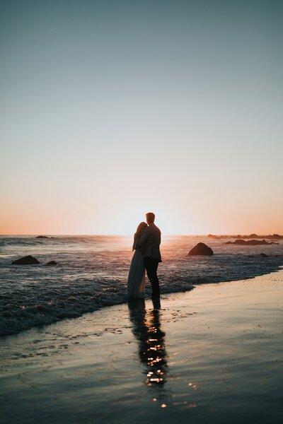 Silhouetted couple embracing at the edge of the ocean during sunset, with gentle waves washing around their feet and the sun casting a warm glow on the water.