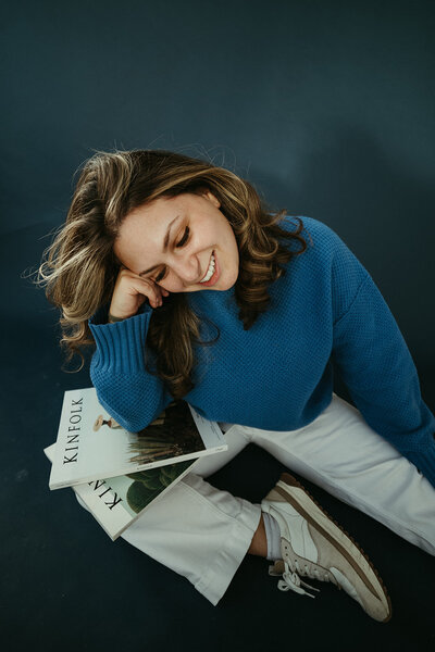 Sara Dobbins sitting on blue backdrop leaning on elbow over magazines