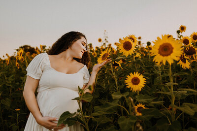 a pregnant woman standing next to a giant sunflower and holding it with her eyes closed in a field in fort worth, texas