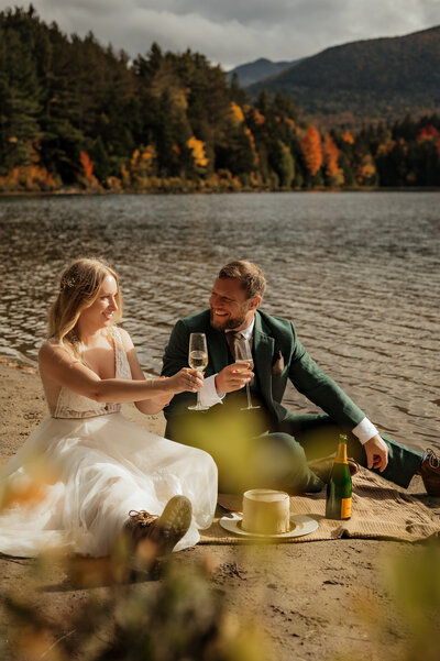 Couple cheers while eating cake by a lake in the Adirondack mountains after getting married. 