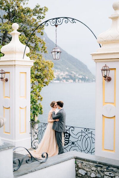 newlyweds-hug-almost-kiss-old-arch-against-backdrop-lake-como-min