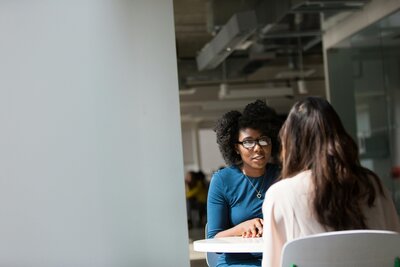 A black woman and a brunette woman sit at a table for an interview. The Burnette's back is turned to the camera.