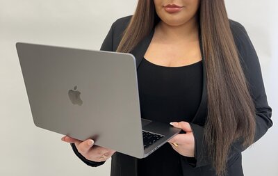  A young woman with light auburn hair wears a dark beige blazer over a smart black cropped bandeau top and high-waisted black trousers. She looks directly at the camera with a poised expression, leaning on a wooden table.