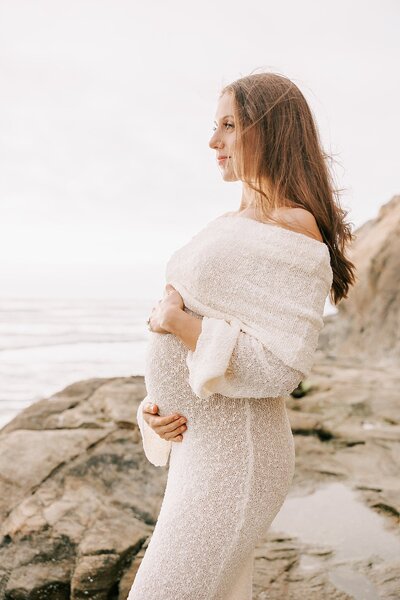 woman holding her belly wile standing on the rocks at the oregon coast. She is pregnant and wearing a white dress off  the  shoulder
