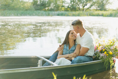 A man and a woman are sitting in a rowboat on a lake. The man is holding his fiance in his arms and she is looking up at him, smiling.