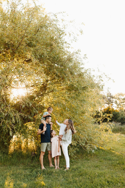 A family of five standing with the baby on dads shoulders all looking at one another and laughing while they're photographed at the park in christchurch by lucy jane photography