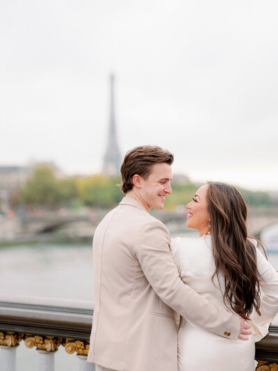 a man in a tan suit and woman in a white dress with their backs towards the camera looking at each other and smiling with the eiffel tower in front of them in the distance
