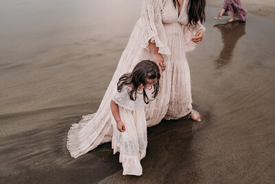 looking down at a mother and her  3 year old daugher looking down at their feet in the sand as they hold hands at the beach. They are both wearing soft pink lace gowns that drape into the wet sand.  The mother's face is not visible.