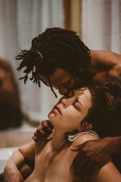 birthing photo of woman looking up while her biracial partner supports her by holding her in the birthing tub and kisses her on the forehead during a contraction