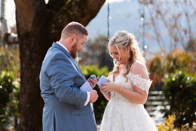 A bride and groom stand outside under a large tree, exchanging heartfelt gifts before their wedding ceremony. The groom, wearing a blue suit, holds a small box while the bride, in an off-the-shoulder lace gown, looks down at her vows with a joyful expression. The soft sunlight and scenic mountain backdrop enhance the romantic moment.