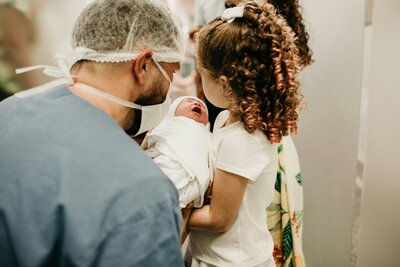 A father holding his newborn while an older sibling looks on with curiosity and tenderness during the early days of adjustment.