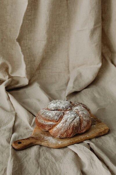 A rustic, golden-brown artisan bread dusted with flour, resting on a wooden cutting board with a linen backdrop, showcasing fresh baked goods from Sleeping Dog Plants.