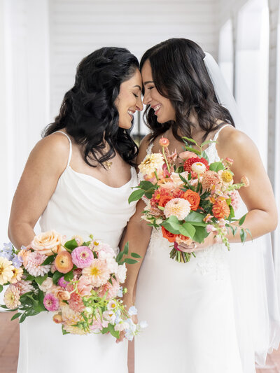 Two brides in white dresses smile and gently touch foreheads, holding vibrant bouquets of orange, pink, and peach flowers. They stand on a porch, celebrating their wedding day with joy and affection.