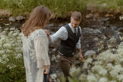 groom leads bride to a river bed amongst tall aildflowers