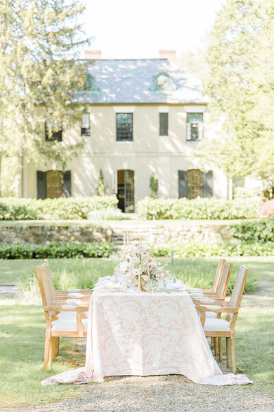 An intimate table setting outdoors in a french-countryside venue with a chateau and gardens in the background. The image featured the table setting with a cream and white damask tablecloth, light wood chairs with white upholstery and white, blush, and cream flowers. Gold candlesticks and flatware. Captured by best destination wedding photographer Lia Rose Weddings