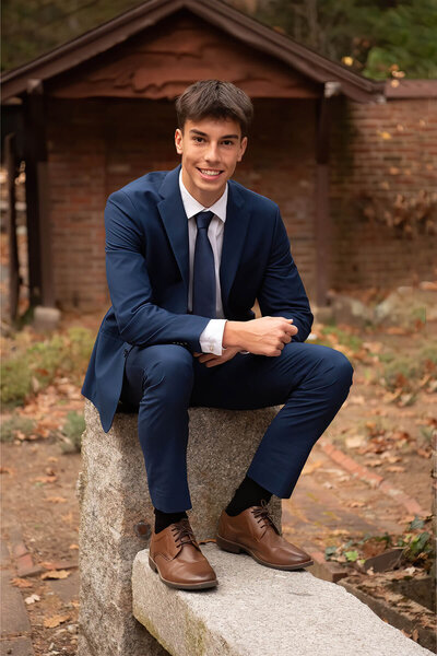 A senior in a navy suit sits during his photo session at Whip Hill Park in Stoneham, MA