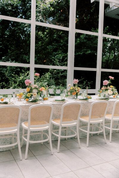 Long table with pastel floral centerpieces and white cane-back chairs inside a bright pavilion with garden views.