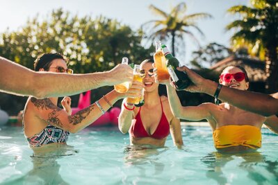 A group of young adults toast their drinks to each other while standing in a blue swimming pool on a  sunny day.
