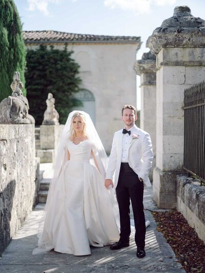 Elegant bride and groom standing hand in hand in a sunlit stone courtyard in the Chateau de Naudou South of France — Portfolio Thomas Raboteur Wedding Photographer.
