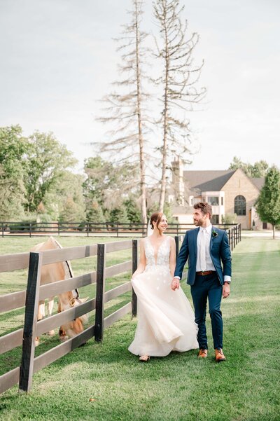 bride and groom walking while holding hands next to fence with horse