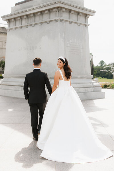 Brunette bride and groom walking toward statue in St. Louis, with bride looking at the camera, by wedding photographer Jacoby Andrick