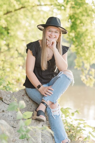 a girl in a black hat sitting on rocks by the water