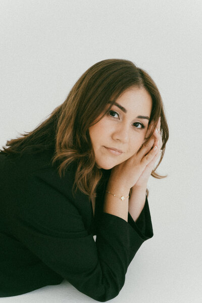 Photographer in black blazer posing on the ground in a clean studio setting with a soft expression.
