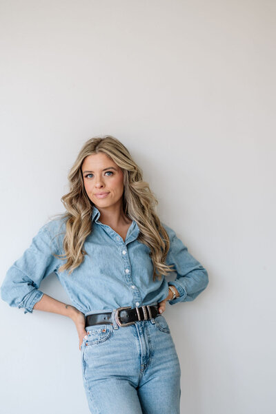Leaning against a white wall, Emily softly smiles with her hands on her hips. She is wearing light wash denim jeans and a match denim button up.