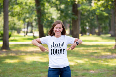 A woman smiling and pointing at her shirt with the words 'Start Today' at a park.