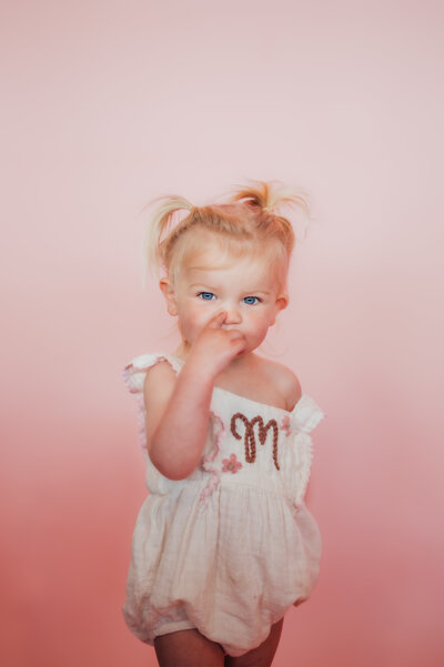 Photograph of 2 year old standing in front of a pink backdrop and sucking her thumb. 