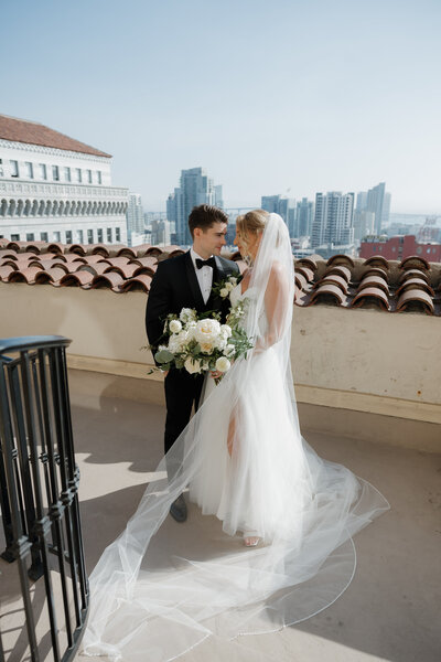 Bride and groom standing on a rooftop with the San Diego skyline in the background, bride wearing a tulle gown and veil, groom in a black tuxedo holding a bouquet.