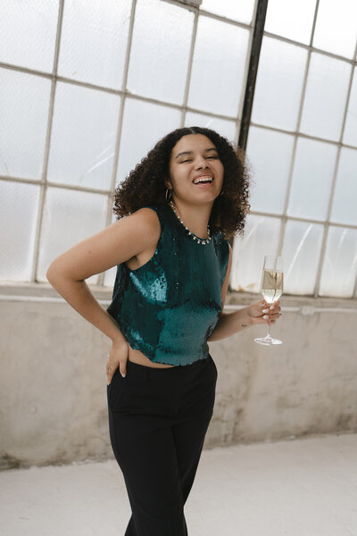 Photo of Catryce holding a champagne class smiling in a teal shirt with a window background
