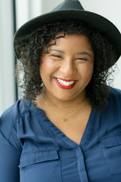 A warm and inviting portrait of Mica McCook, an Austin-based food photographer known for her storytelling visuals. In this image, Mica is smiling brightly, wearing a black hat and a dark top, capturing her approachable and creative personality.