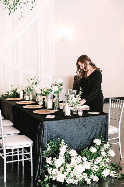 A woman arranges floral centerpieces on a black velvet tablescape at SoHo63, a Chandler wedding and event venue. The table is set with gold chargers, candles, and lush white florals, creating a refined and modern look for an intimate event or reception.