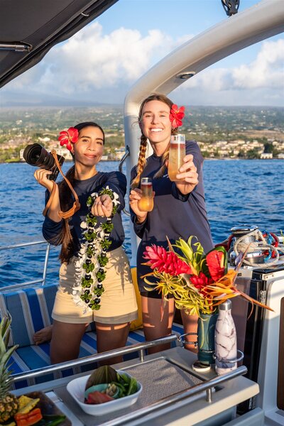 Sailboat crew in Hawaii smiling and holding drinks and a camera, celebrating during a sunset proposal charter with tropical flowers in the background.