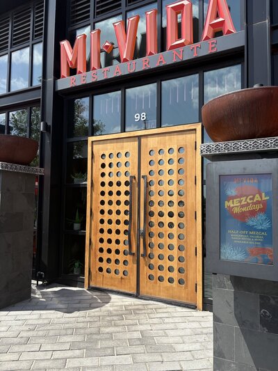 Exterior of Mi Vida Restaurant at the Wharf in Washington, D.C., featuring old red signage and large wooden double doors with circular cutouts, framed by black trim and modern architectural details. 