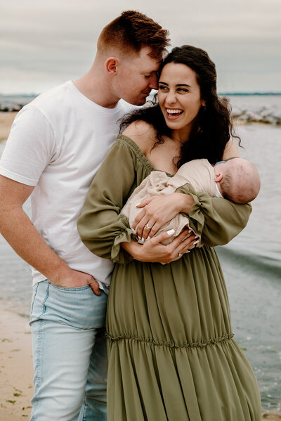 Dad in white shirt and jeans whispering to mom in green dress while she is laughing and holding baby boy in Severna Park Maryland.