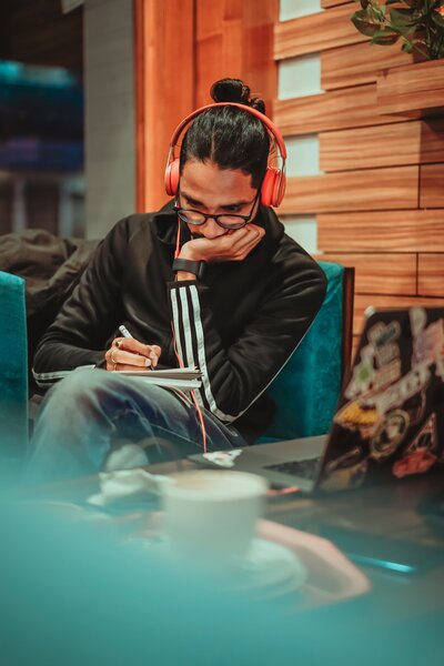 A brown-skinned man in a black track jacket sitting on a turquoise couch. He is wearing orange headphones and writing in a notebook. Photo by Dollar Gill via Unsplash.