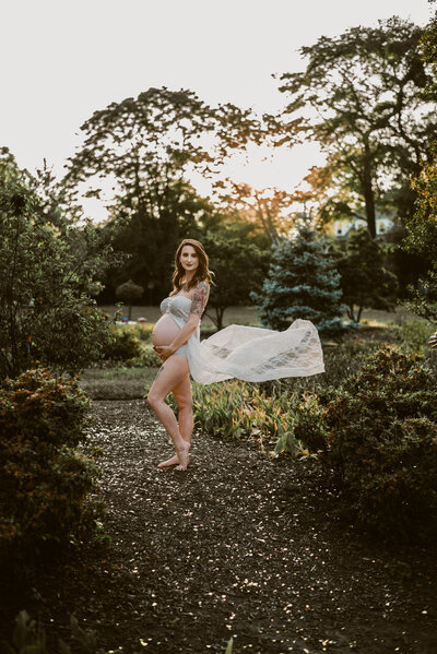 Pregnant mom in white dress flowing behind her in garden with beautiful sun setting behind her in Baltimore Maryland.