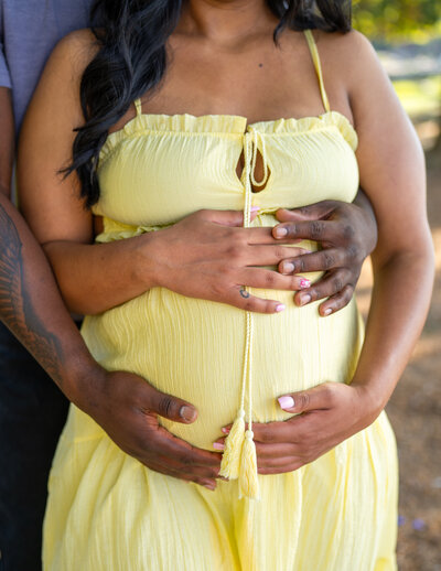 A father and mother's arms cradle the top and bottom of a her belly over her pale, yellow sun dress. Photo by SAVI Photography - Photographer in Riverside